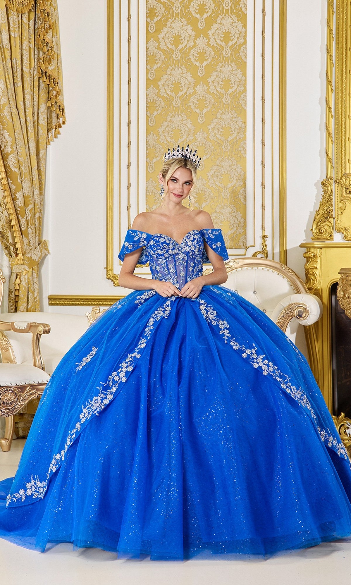 A woman in the Quinceanera Dress 5066 By Dancing Queen, featuring royal blue fabric and silver embroidery, wears a tiara while standing in an ornate room with gold accents and classic decor.